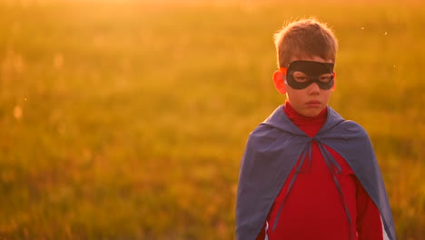 A-child-in-the-costume-of-a-superhero-in-a-red-cloak-runs-across-the-green-lawn-against-the-backdrop-of-a-sunset-toward-the-camera.